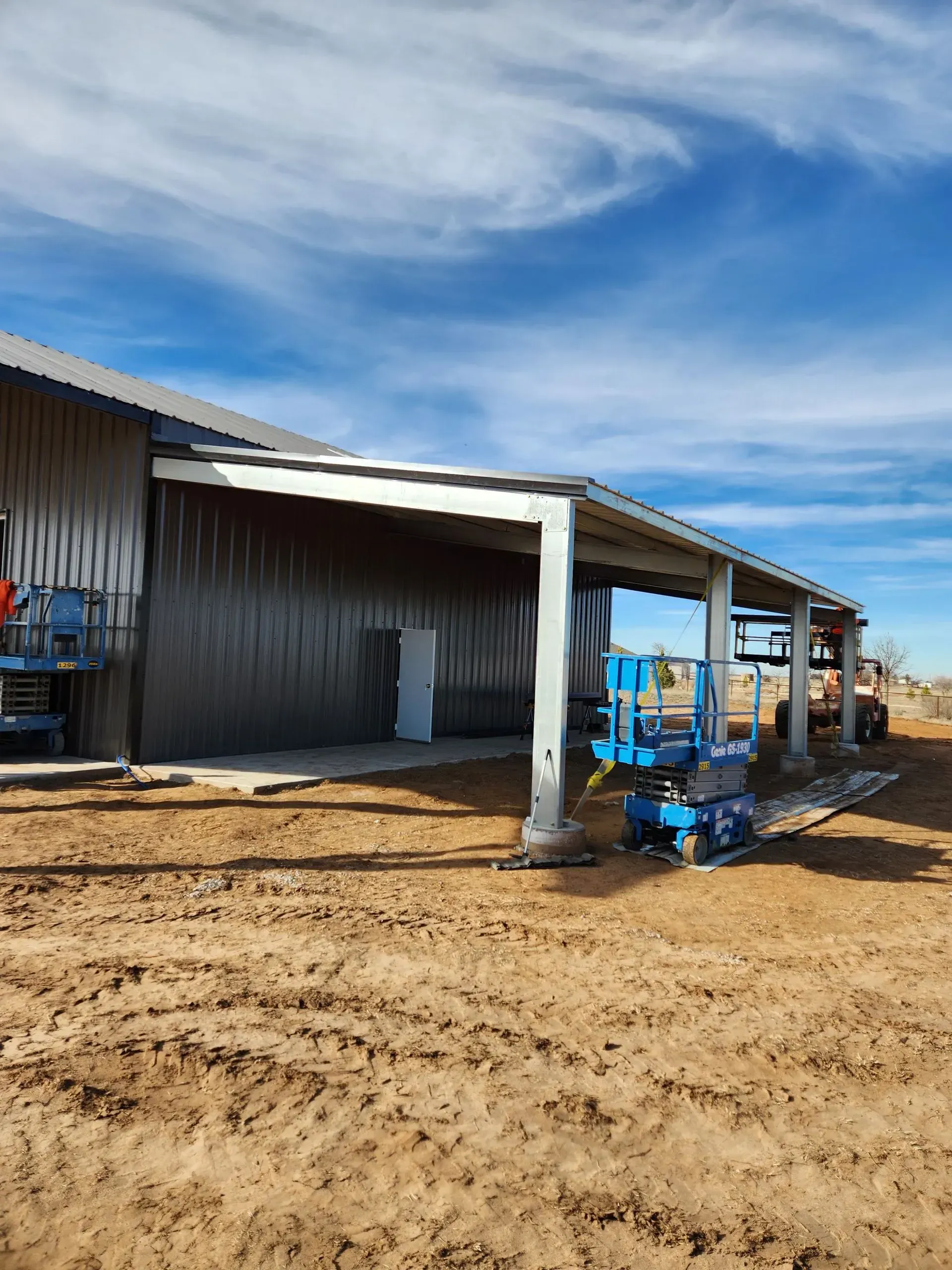 Blue farm equipment parked beside a metal barn on a muddy dirt lot under a cloudy sky
