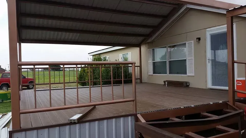 Covered wooden porch attached to a beige mobile home, with stairs and a fenced yard.