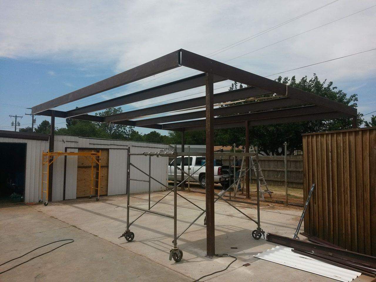 Steel canopy frame under construction in a yard, with scaffolding and wooden fencing nearby