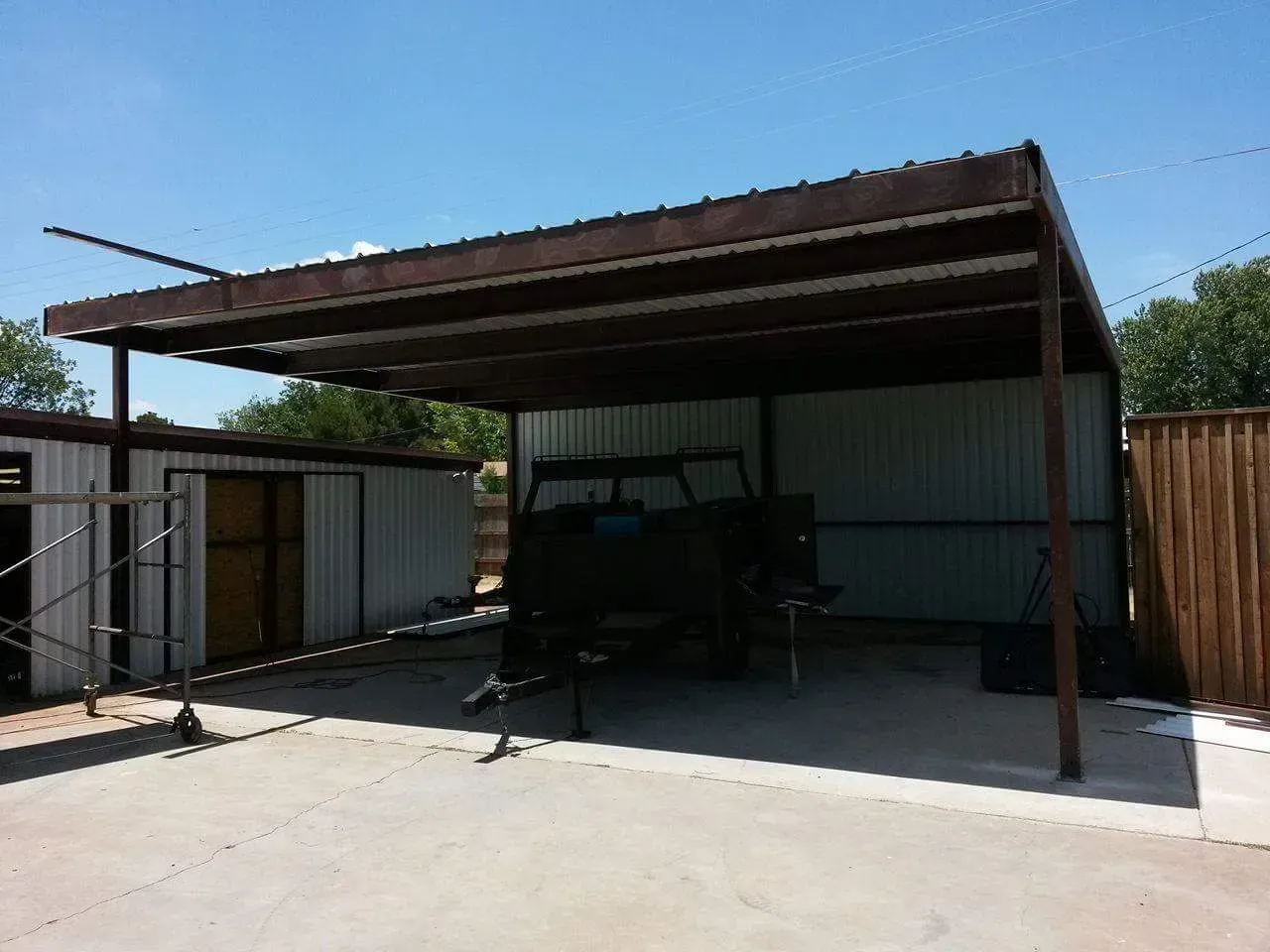 Carport with a parked vehicle under a metal roof beside a white building and wooden fence