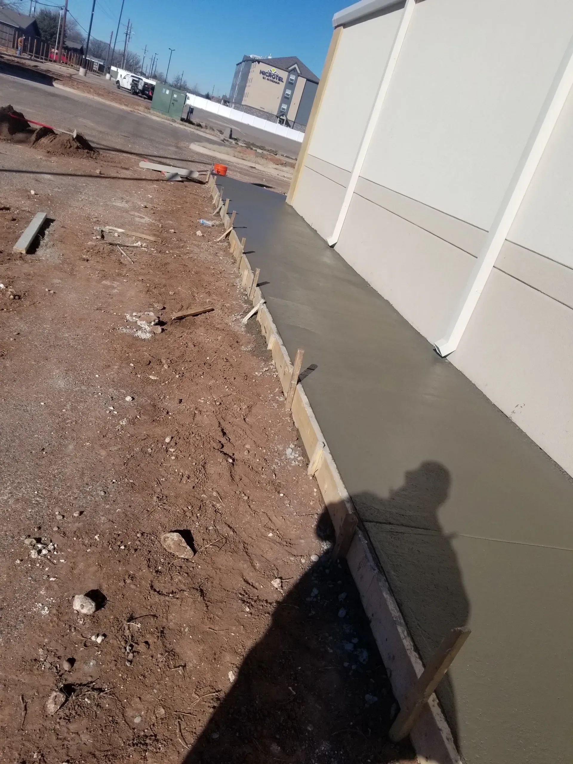 Construction site beside a white wall with a dirt trench and wet concrete walkway in bright sun