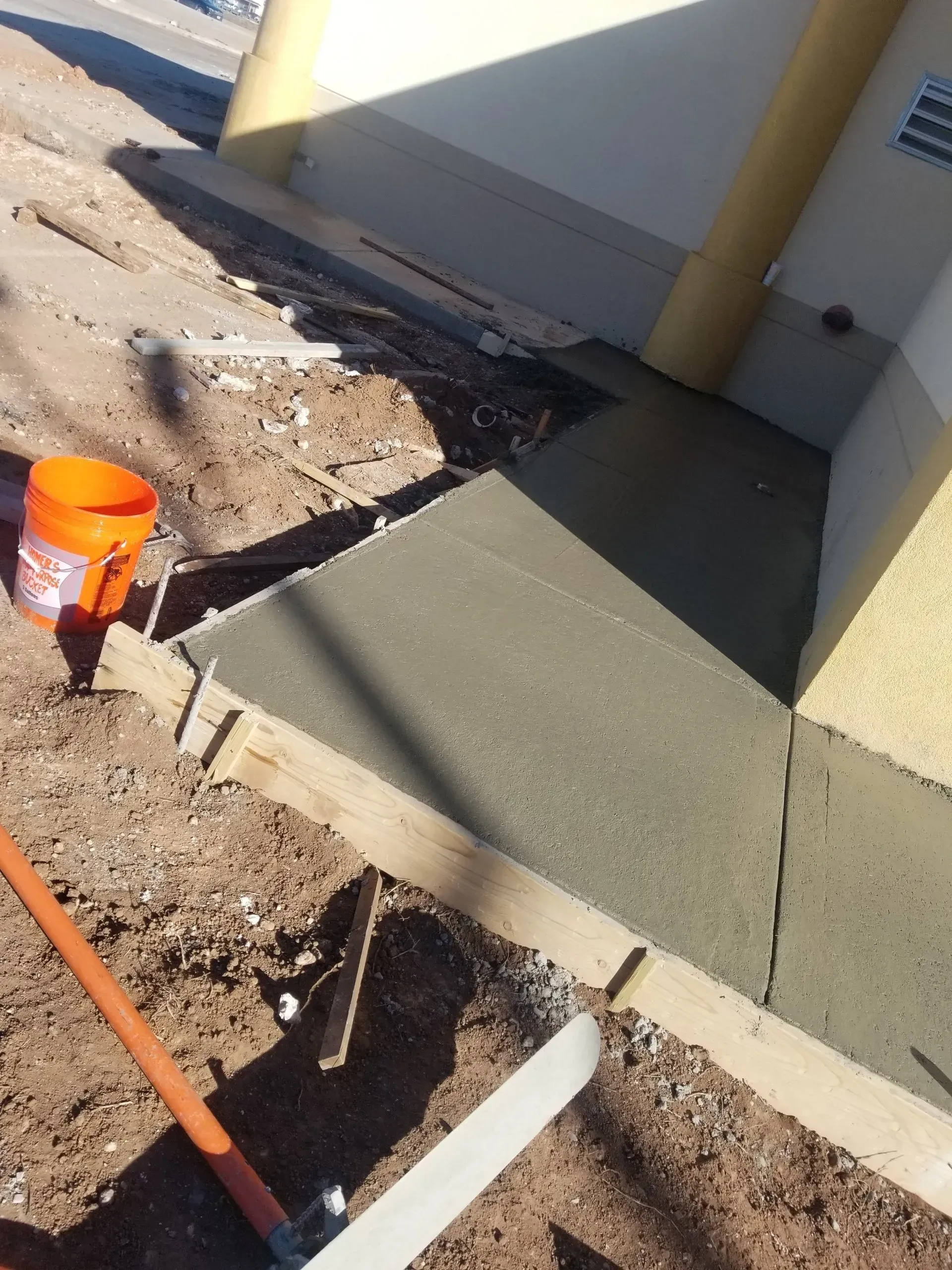 Fresh concrete sidewalk ramp beside a building, with dirt trench, orange bucket, and shovel in foreground