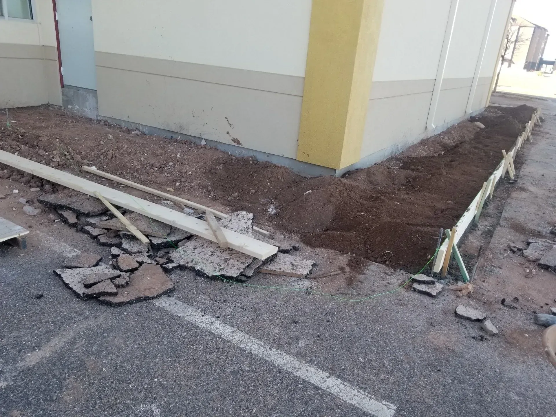 Damaged sidewalk and broken debris beside a building corner with exposed dirt and a cracked curb line