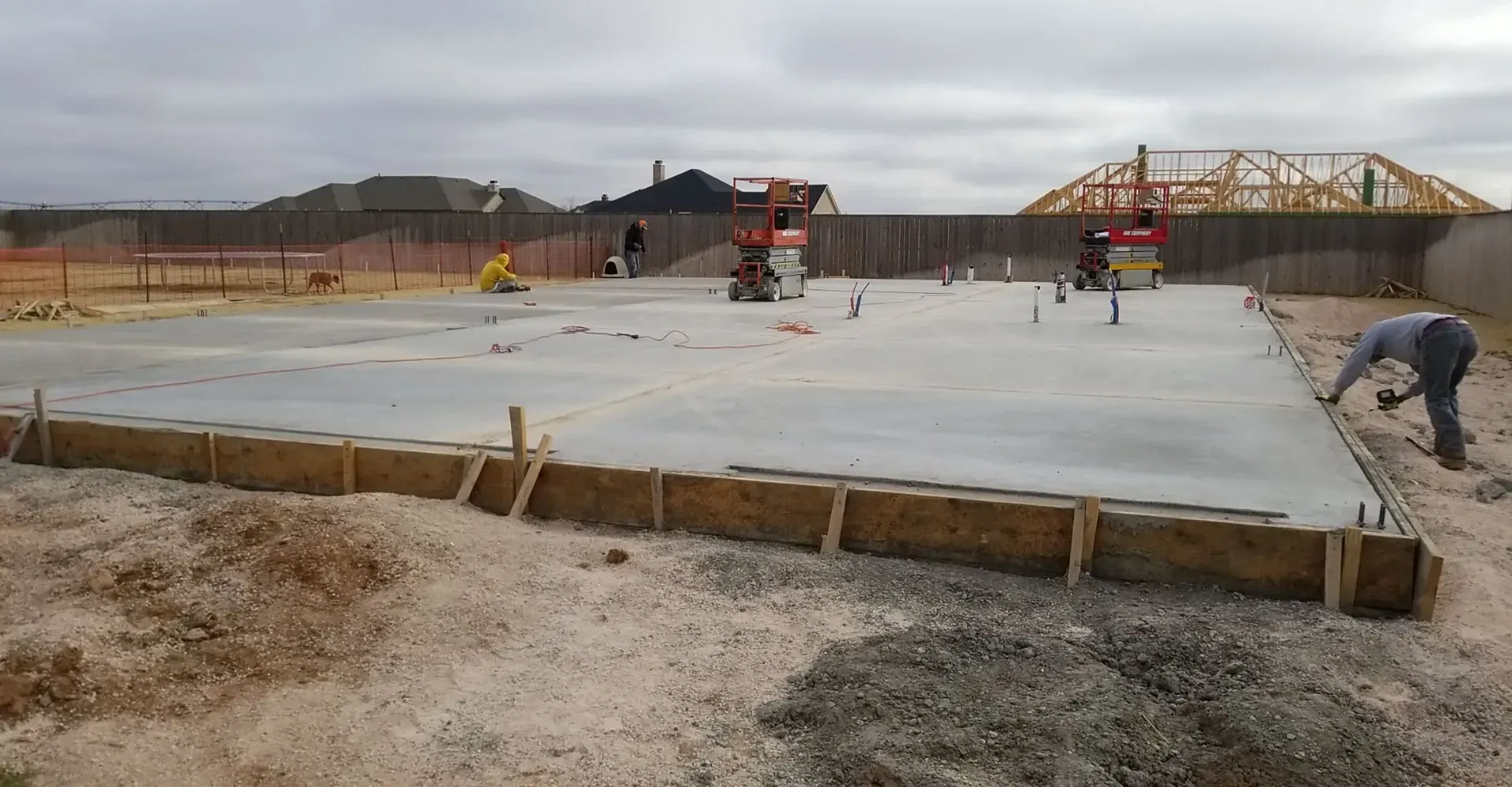 Concrete slab foundation under construction with workers and wooden formwork in a dirt lot