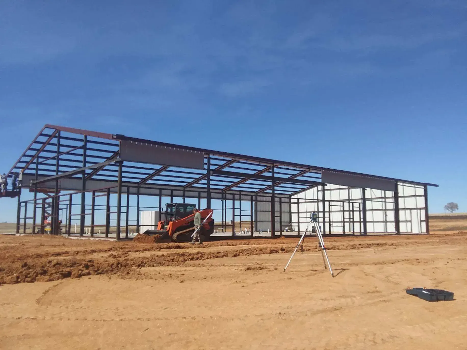 Steel warehouse under construction on a dirt lot, with workers and equipment beneath a blue sky