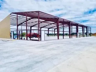 Large red steel warehouse frame under construction on a concrete lot, with vehicles nearby and blue sky overhead