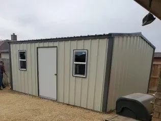 Gray metal storage shed with two windows and a door on a gravel lot