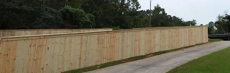 Long wooden fence along a driveway with trees in the background. Gray sky, grassy lawn on the right.