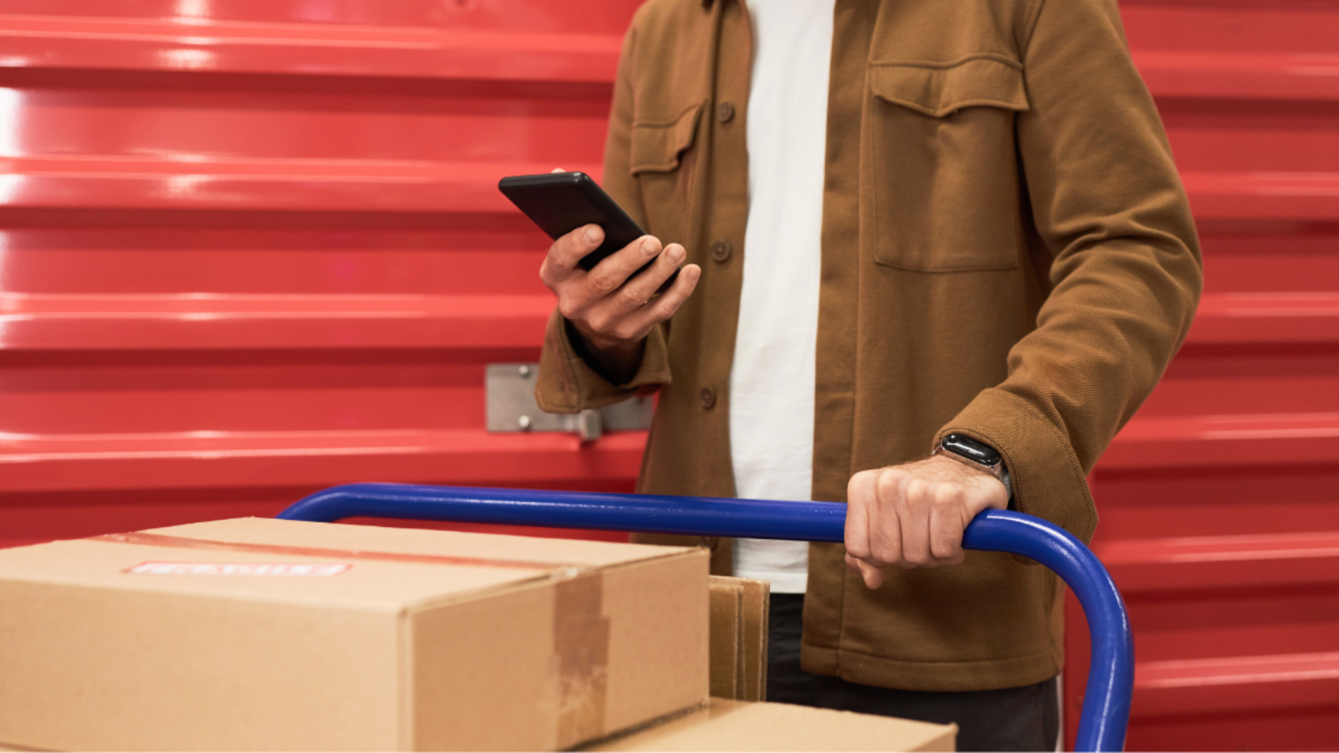 A person in a brown jacket uses a phone while pushing a cart with cardboard boxes in front of a red storage unit door.