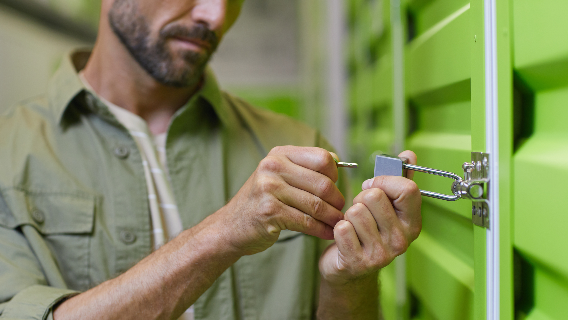 A person in a green shirt unlocks a padlock on a green metal storage unit door.