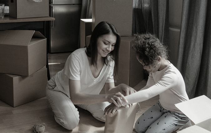 Woman and child on the floor, unpacking items from a box in a room with moving boxes.