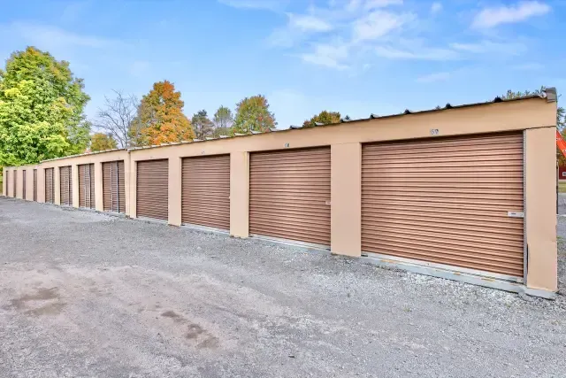 Row of tan storage unit garage doors beside a gravel lot under a blue sky