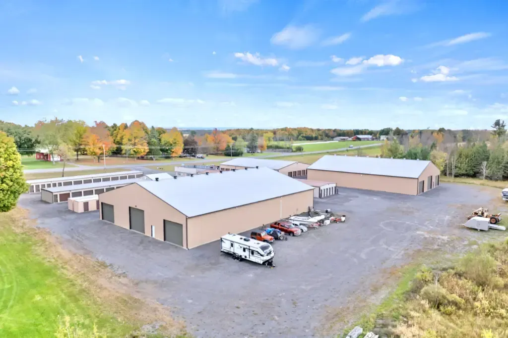 Aerial view of two tan warehouse buildings, trucks, and a gravel lot surrounded by trees under a blue sky.