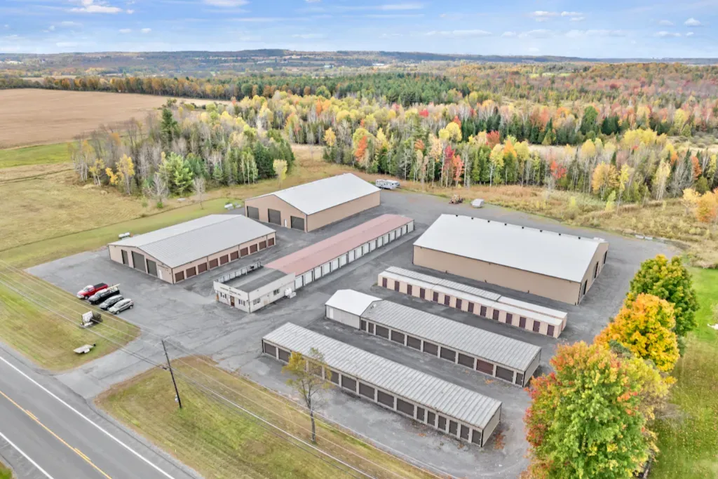 Aerial view of a storage facility with rows of buildings beside a road and autumn trees