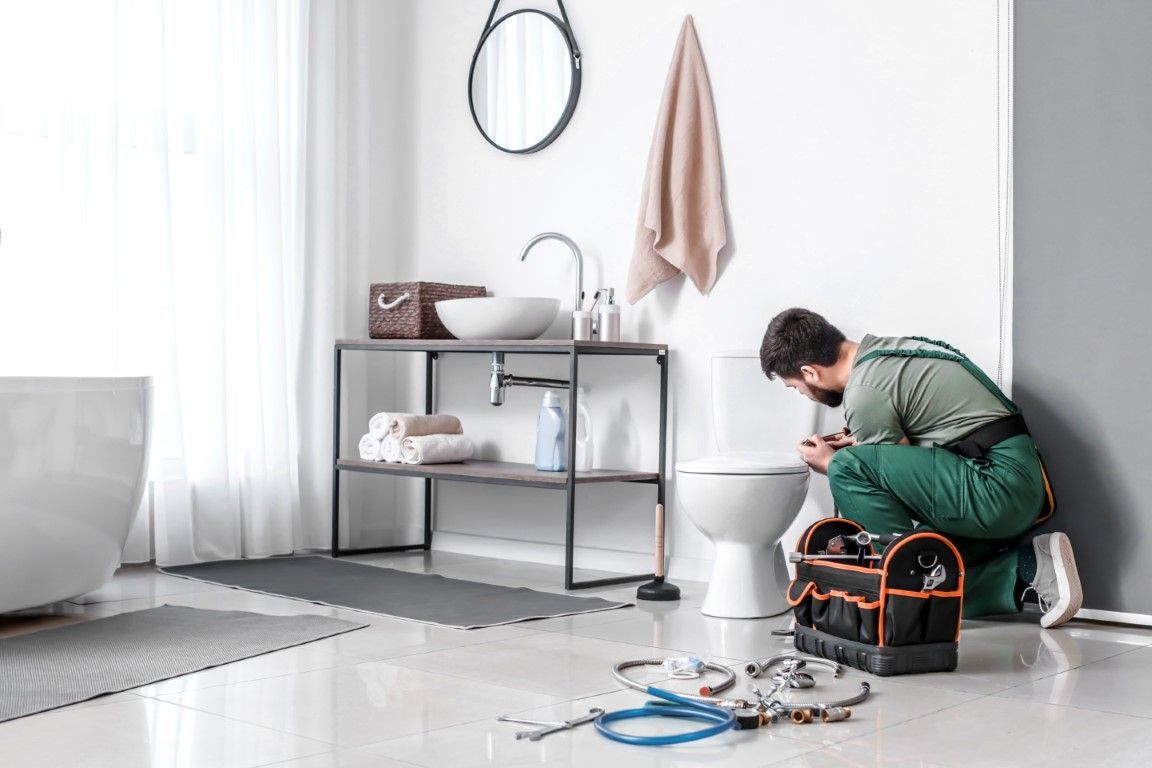 Plumber kneeling near toilet in bathroom; toolbox, tools, towels, and sink visible.