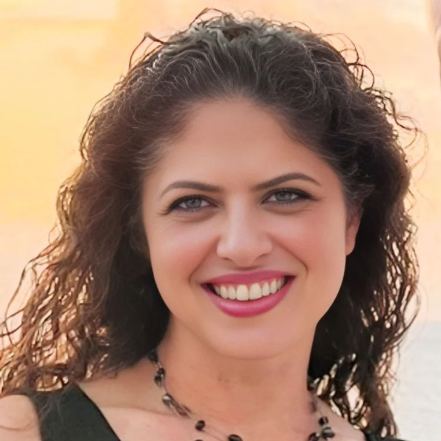 Smiling woman with curly dark hair, wearing a necklace against a warm, blurred background