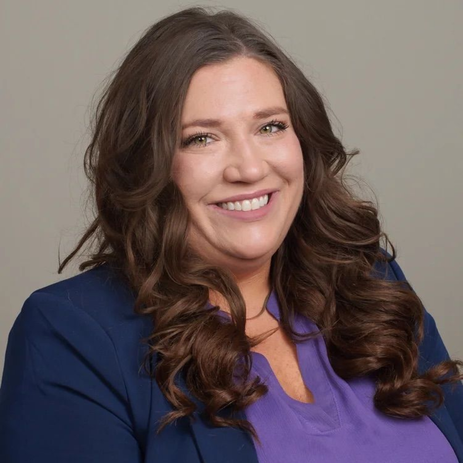 Smiling woman with long brown hair in a blue blazer and purple top against a neutral background
