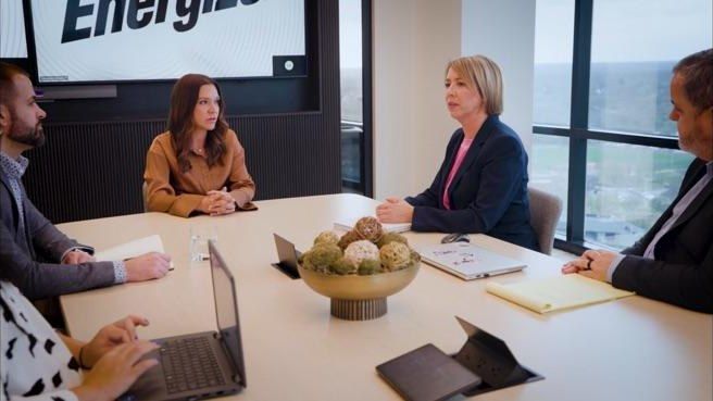 Business meeting around a conference table with laptops, notebooks, and a bowl of fruit near a window