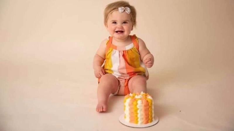 Smiling baby in orange-striped outfit sitting beside a small frosted cake on a beige background