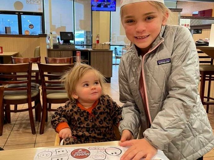 Two children at a restaurant table, smiling beside a menu in a bright dining area.