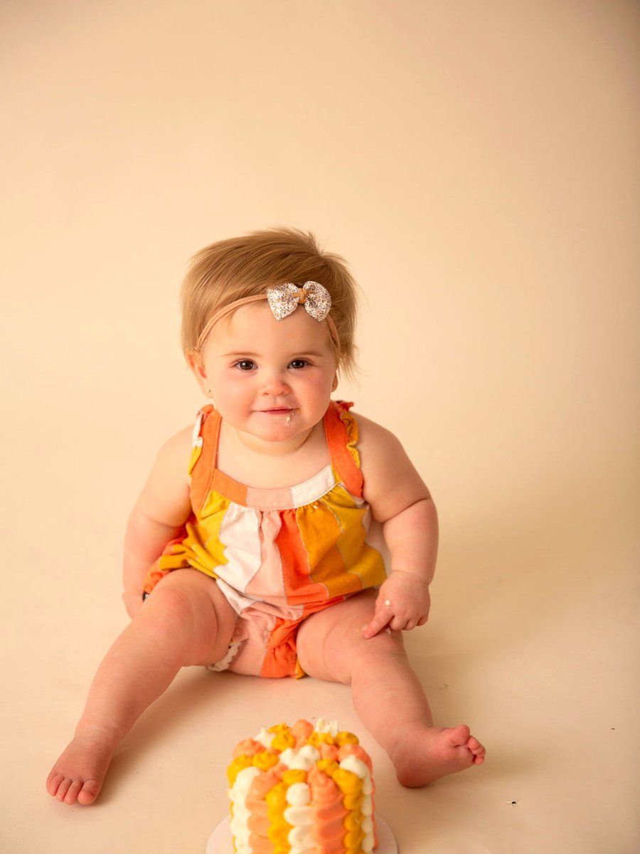 Smiling baby in orange-and-yellow outfit sitting on a beige background beside a small decorated cake