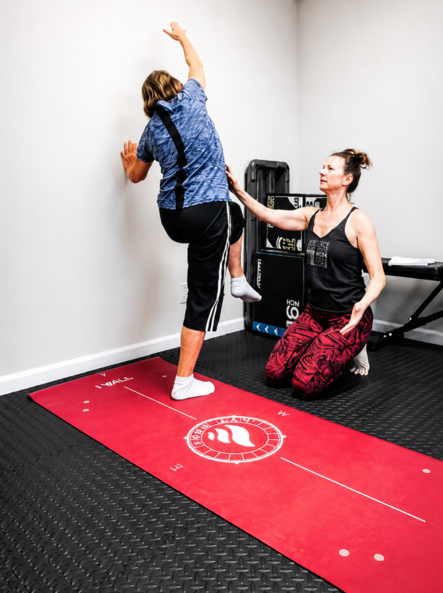 Joi guiding a client through a wall-supported balance and reaching exercise.