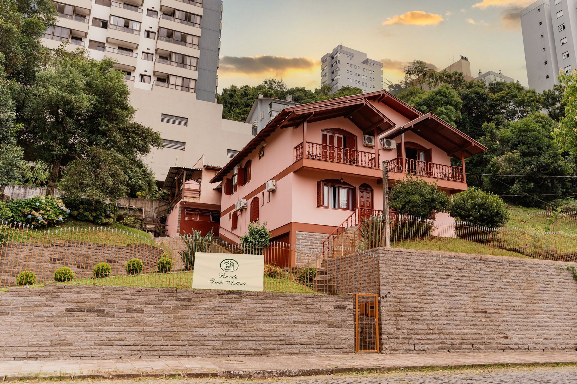 Uma grande casa rosa está situada no topo de uma colina cercada por árvores.- Pousada Santo Antônio