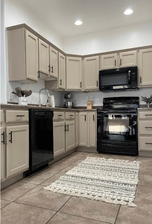 Beige kitchen cabinets with black appliances and hardware, tile floor, small rug.
