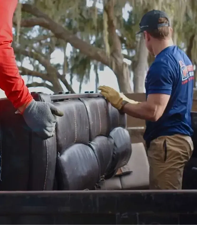 Two workers loading a dark couch onto a truck outdoors, one in red and one in blue.