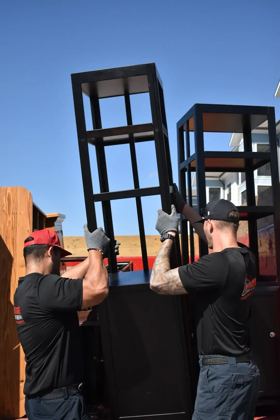 Two workers lift black metal frames outdoors under a blue sky.