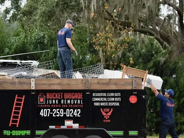 Two workers load debris into a black junk removal dumpster under a tree canopy.