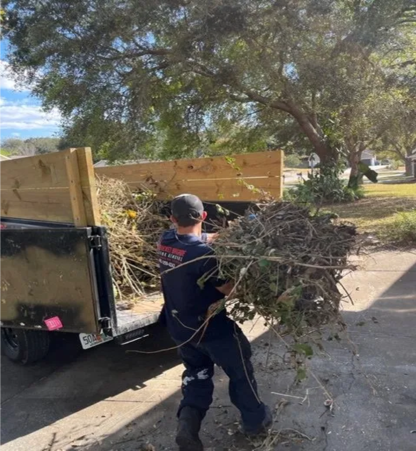 Worker hauling tree branches into a trailer on a sunny street with trees overhead
