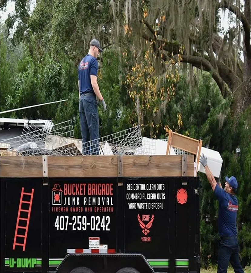 Worker on a trailer roof beside a ladder truck, with a “Bucket Brigade” sign and debris in the background.