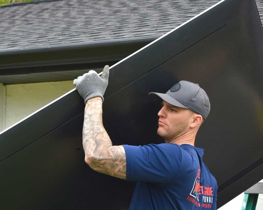 Worker lifting a large black panel outdoors, wearing a cap and gloves.
