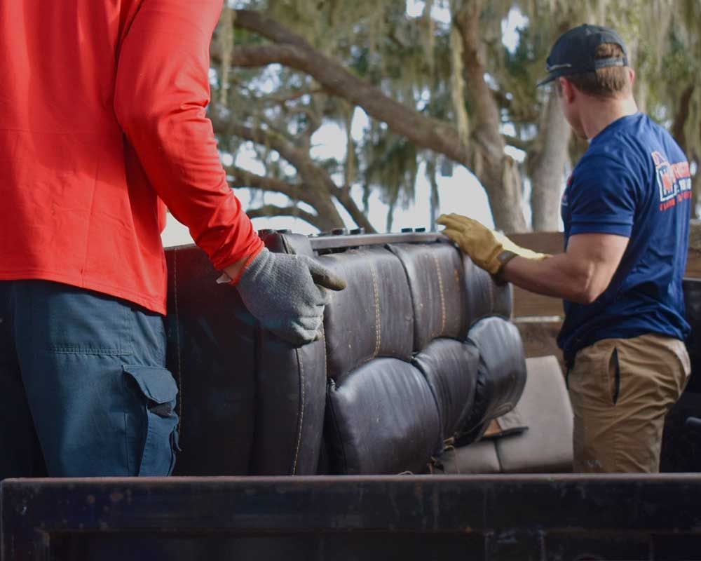 Two workers in gloves lifting a rolled gray couch onto a truck bed outdoors.
