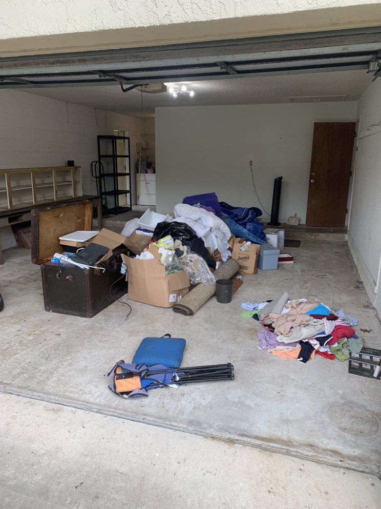 Cluttered garage with boxes, bags, clothes, and tools scattered across the floor.