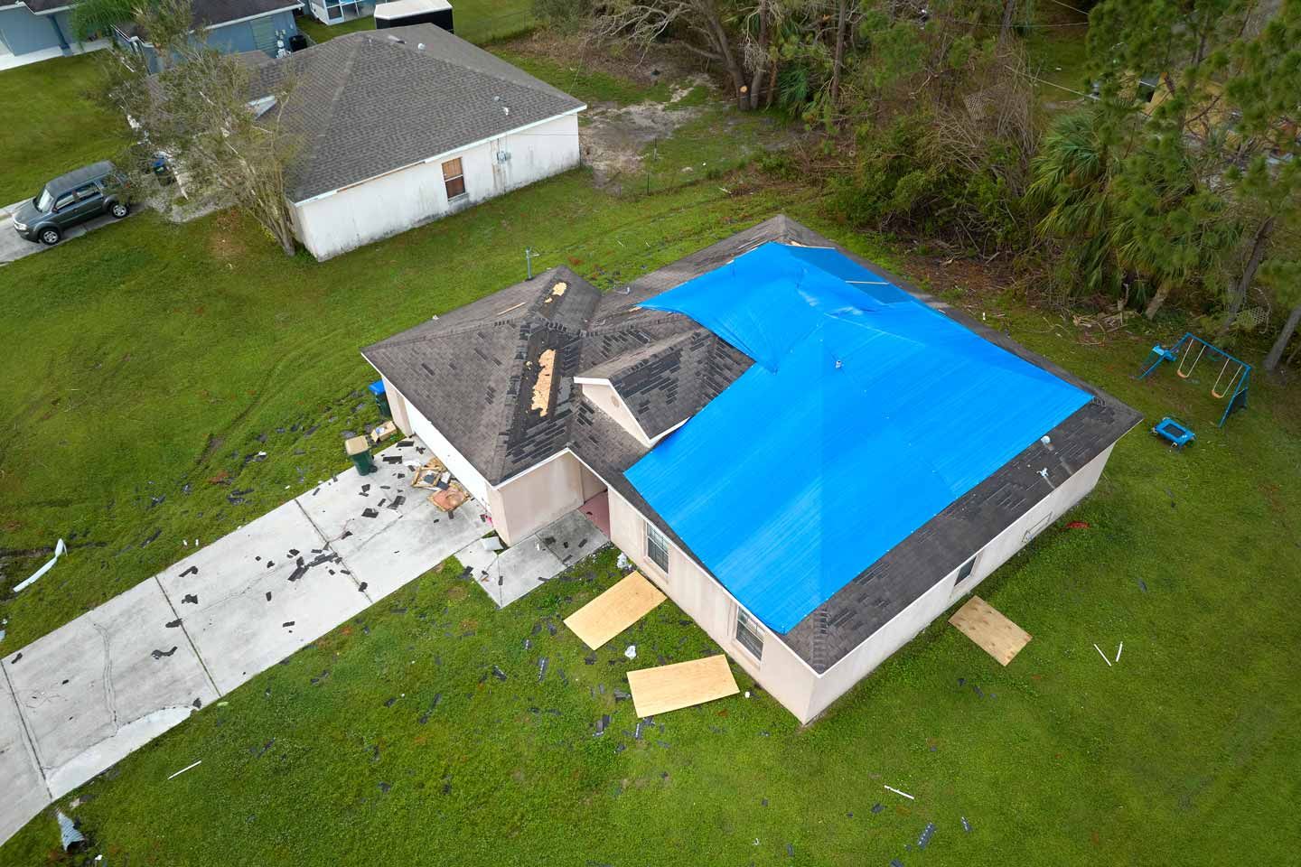 Aerial view of a house with a blue tarp-covered roof and a driveway in a grassy neighborhood.