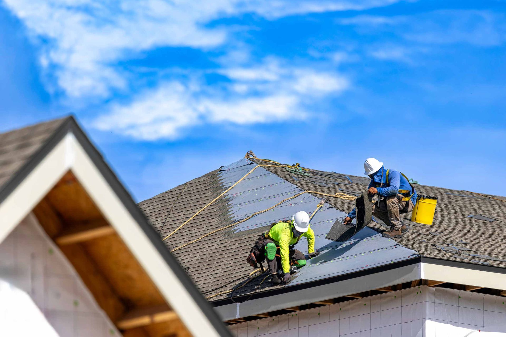 Workers repairing a shingle roof under a blue sky, with bright safety gear and tools on the roof