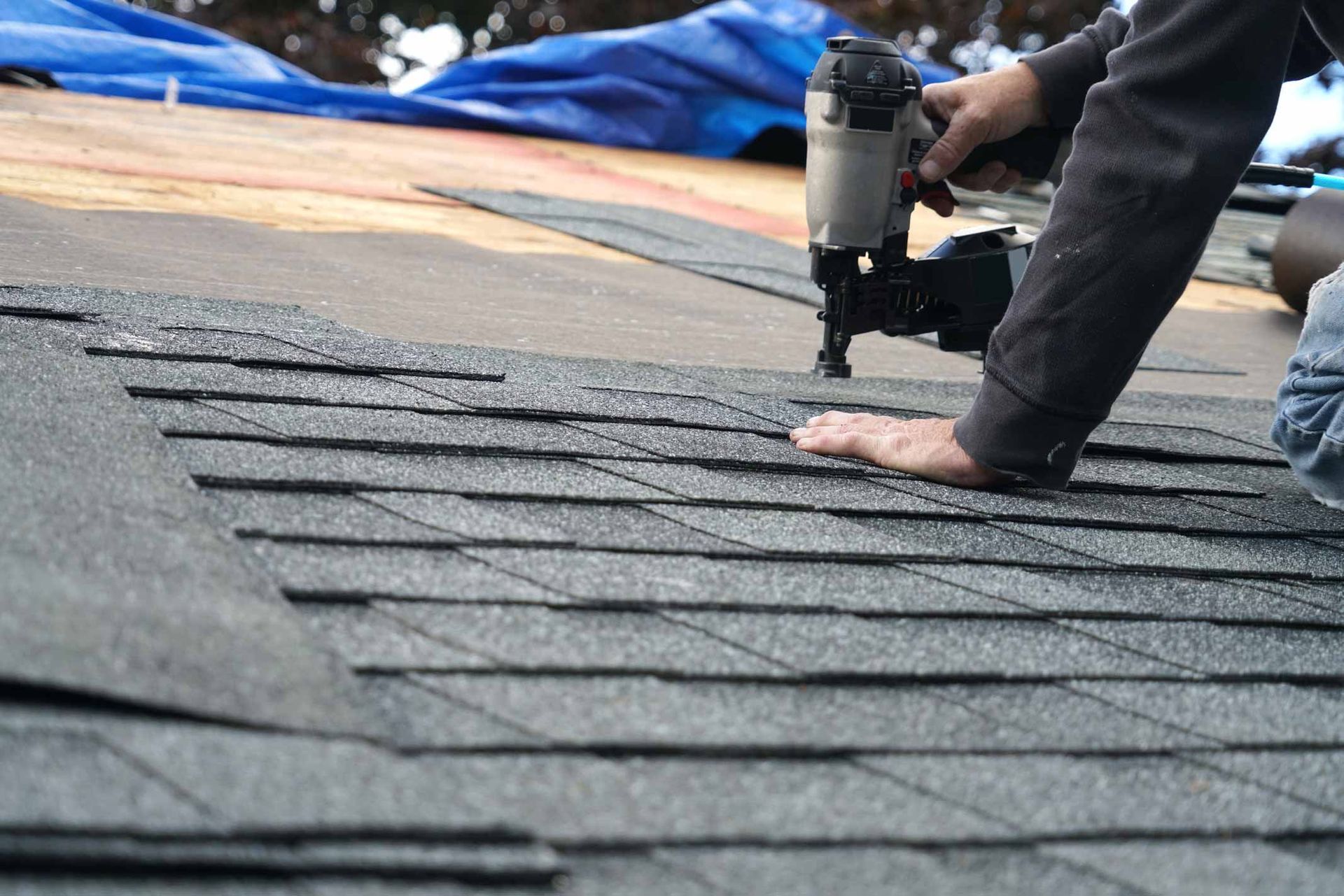 Worker using a nail gun to install dark roof shingles on a rooftop