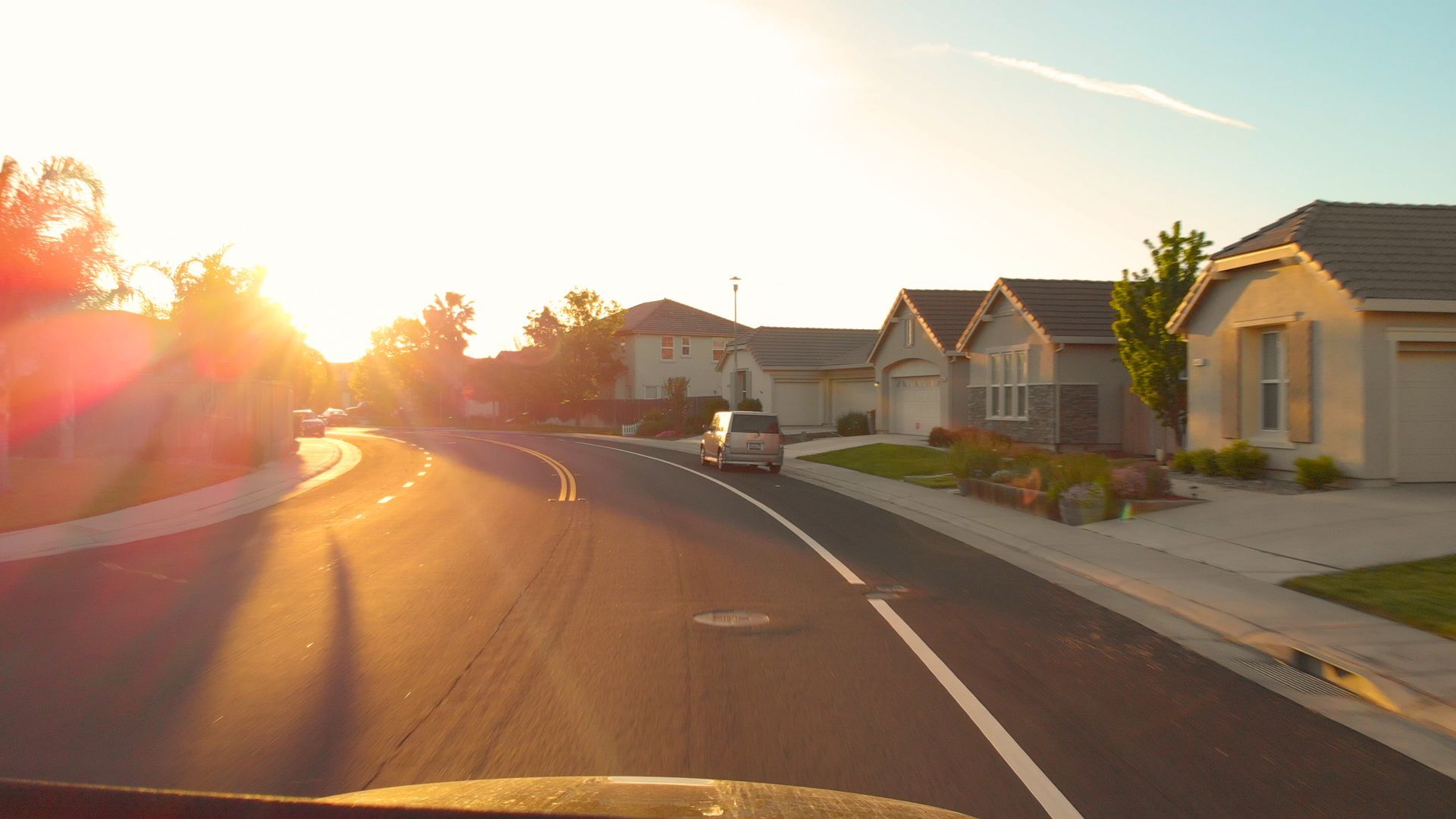 Sunlit suburban street with houses, sidewalks, and a car driving at sunset