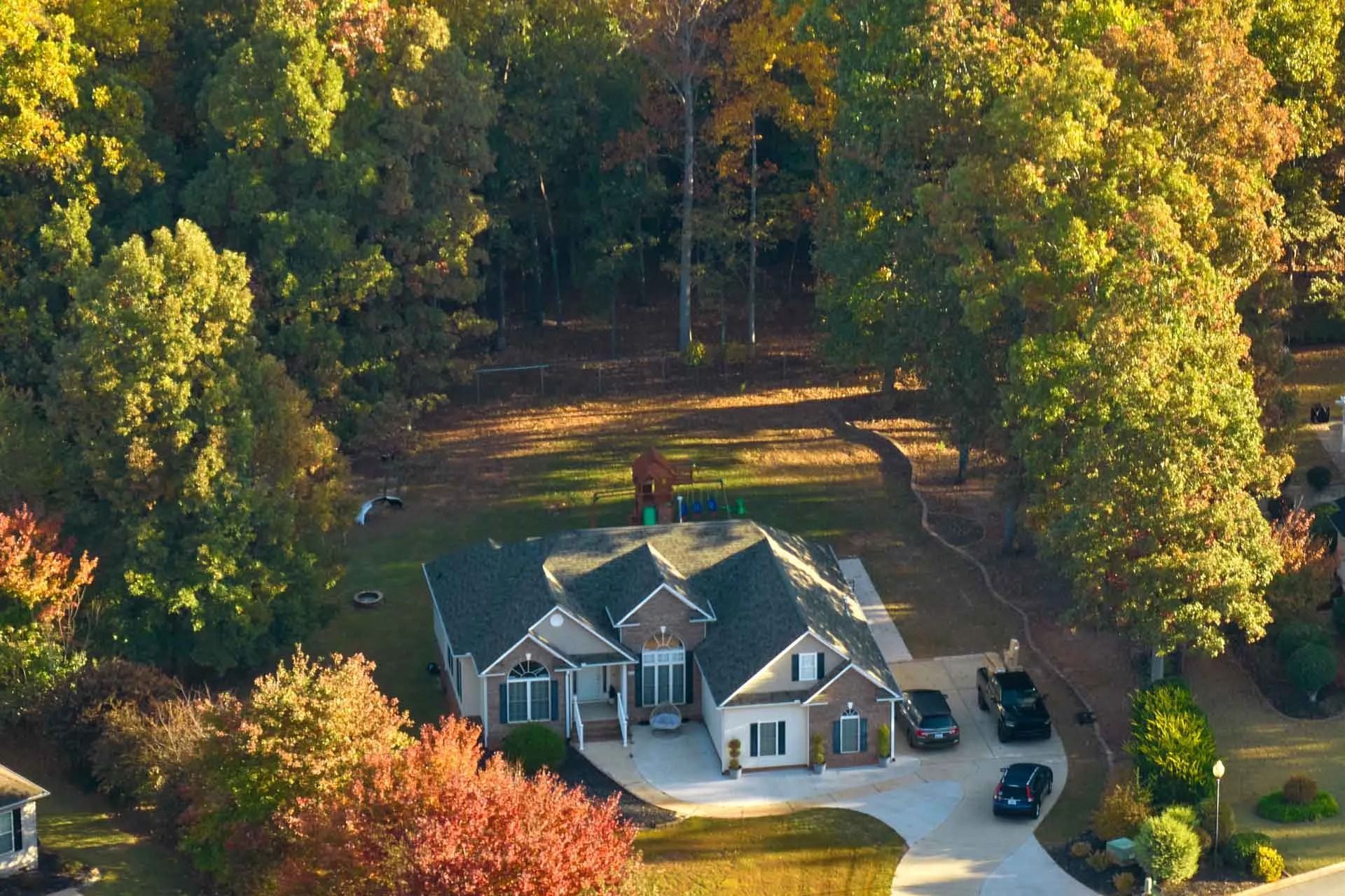Aerial view of a gray house with a driveway, surrounded by trees in autumn colors