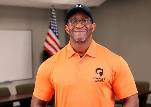 Smiling man in an orange polo and black cap standing in a meeting room with an American flag.