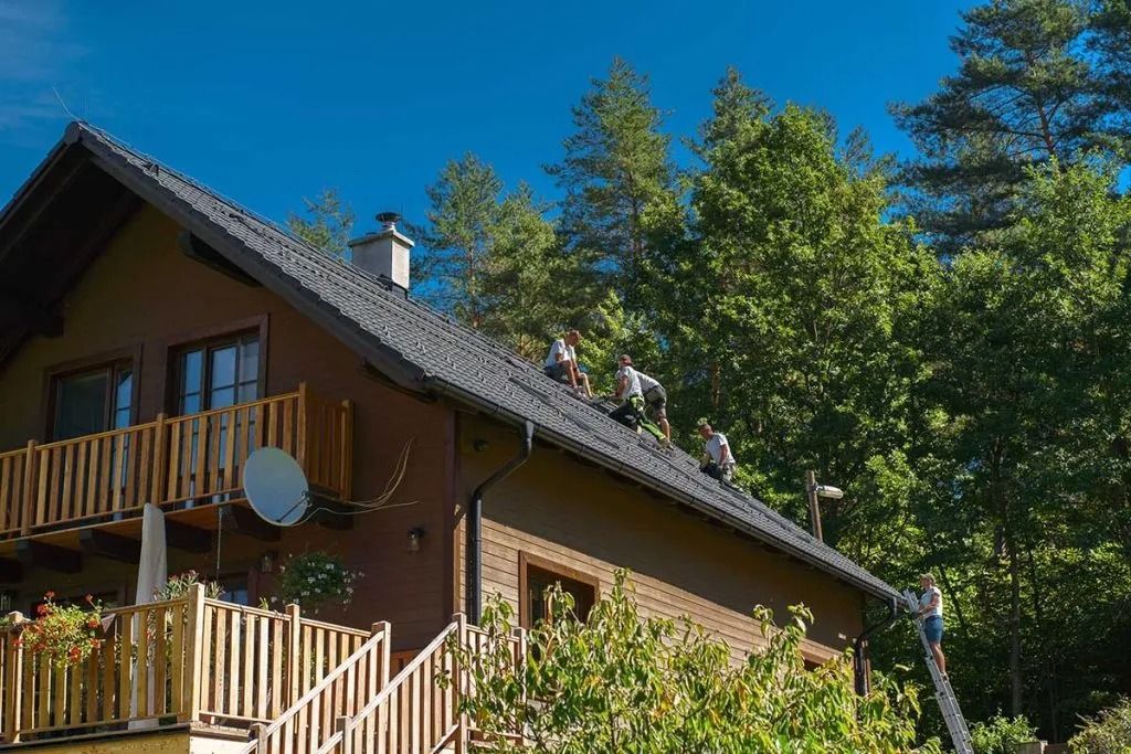 Wooden house with balcony and satellite dish under a bright blue sky, surrounded by trees