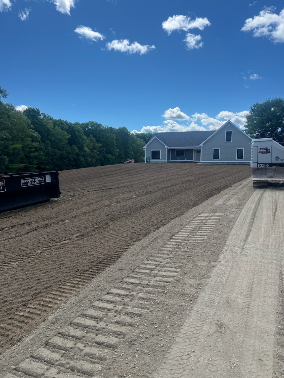 A newly graded dirt yard leads toward a gray suburban house under a blue, cloudy sky, next to a gravel path and dumpster.