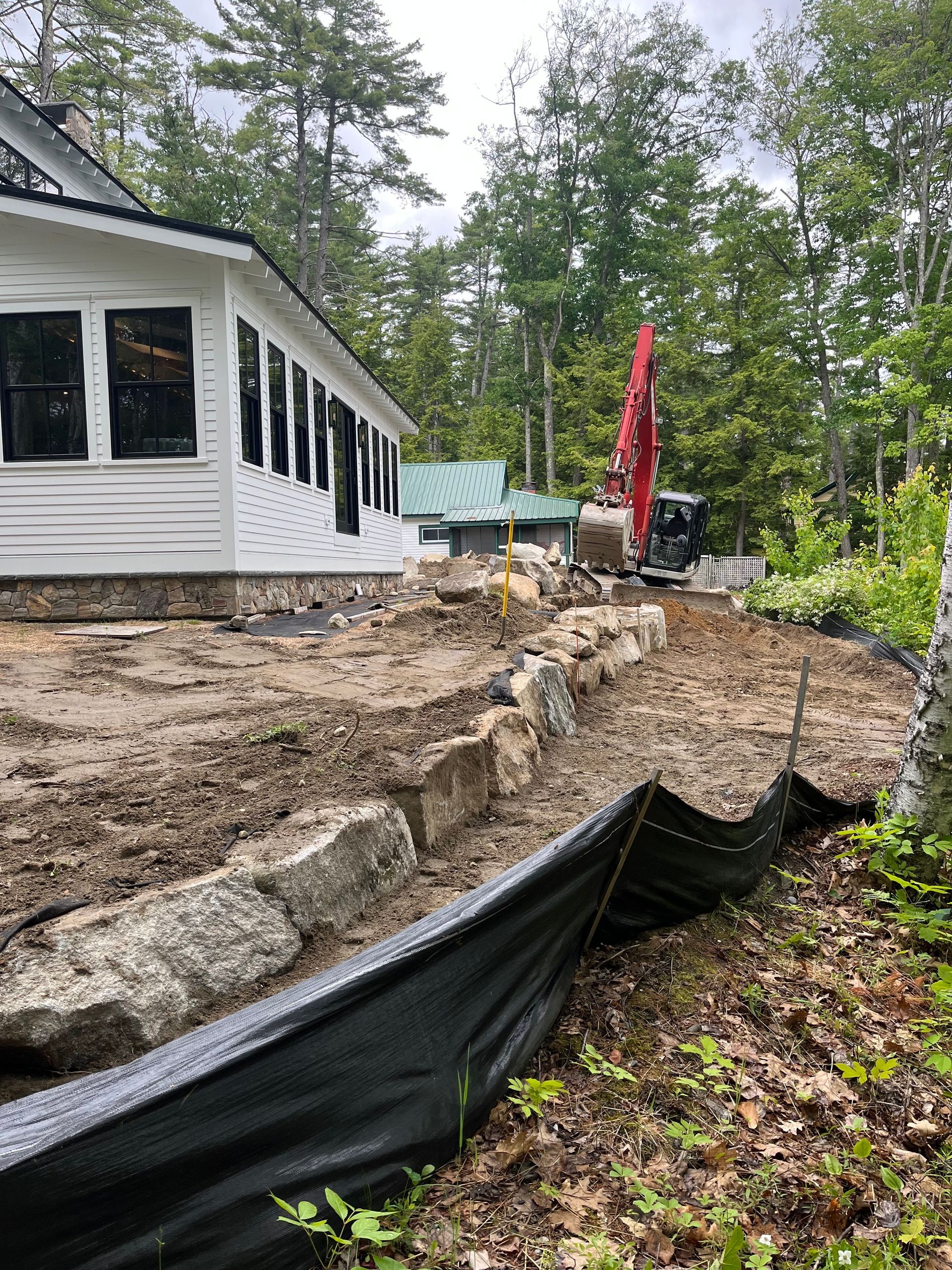 A red excavator parked near a white house under construction, with a stone retaining wall and silt fence in the foreground.