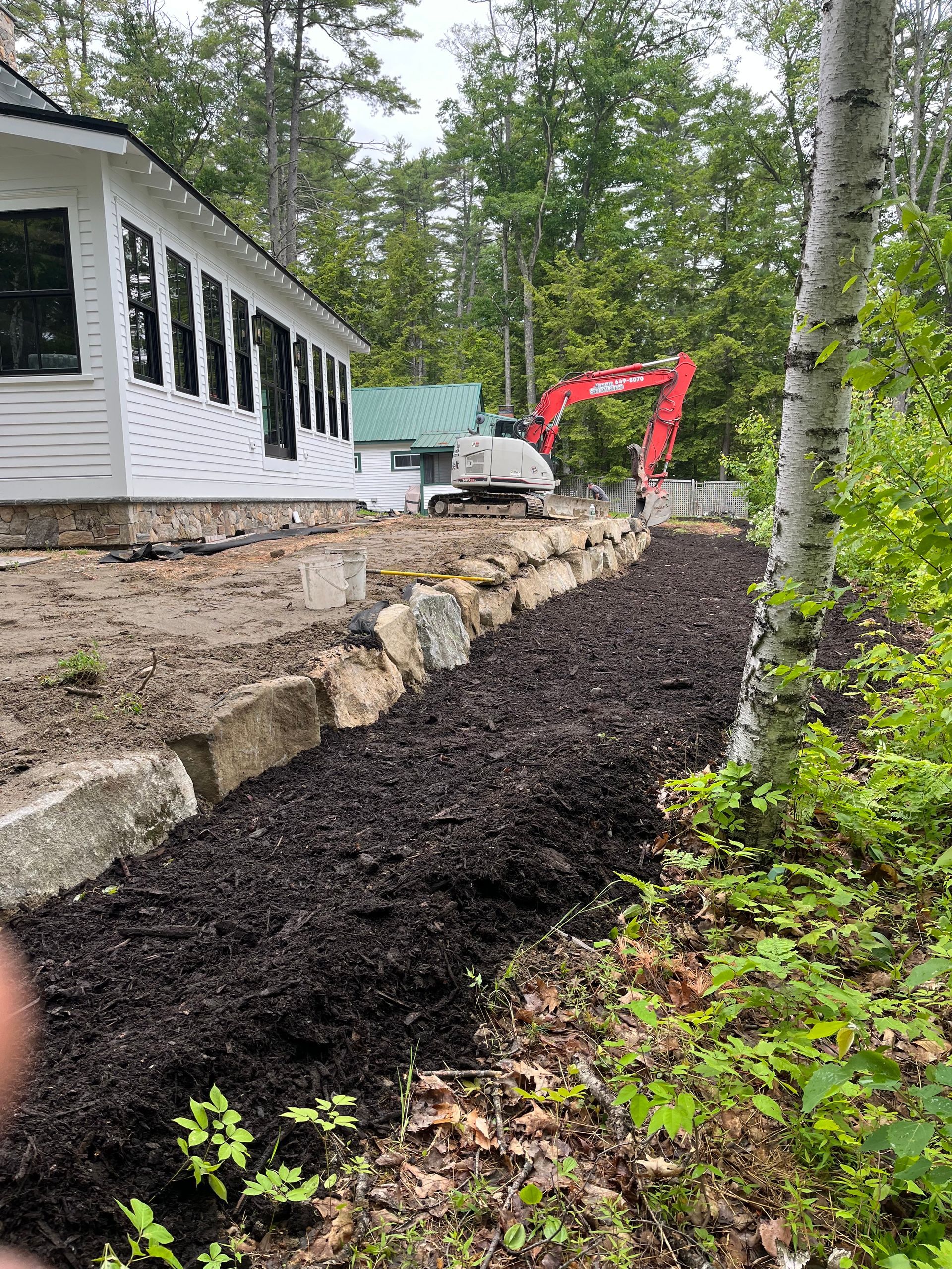 A red excavator parked near a house with a stone retaining wall and a long, fresh bed of dark mulch in a wooded yard.