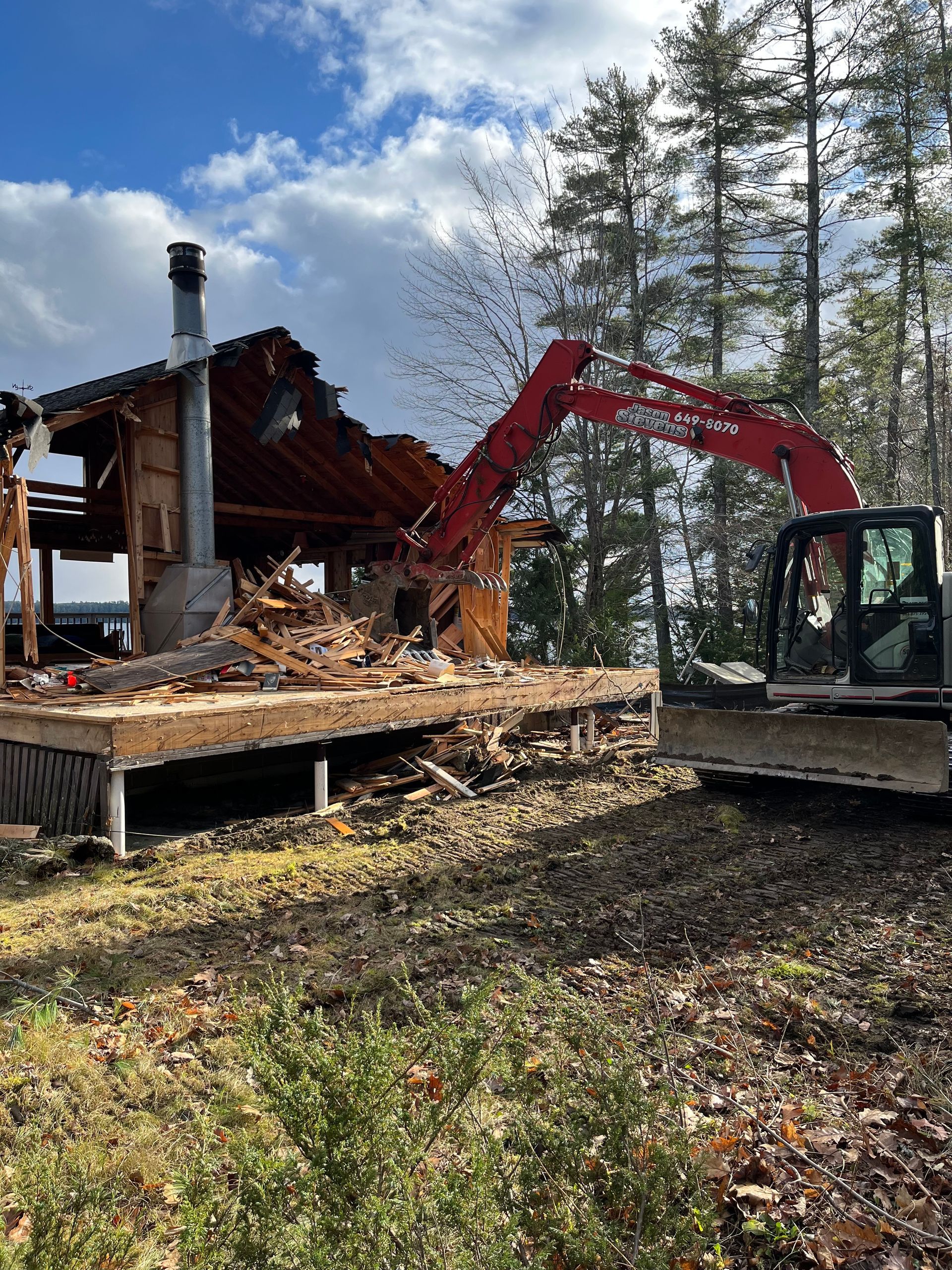 A red excavator demolishing a wooden structure with a prominent chimney against a backdrop of trees and a blue sky.