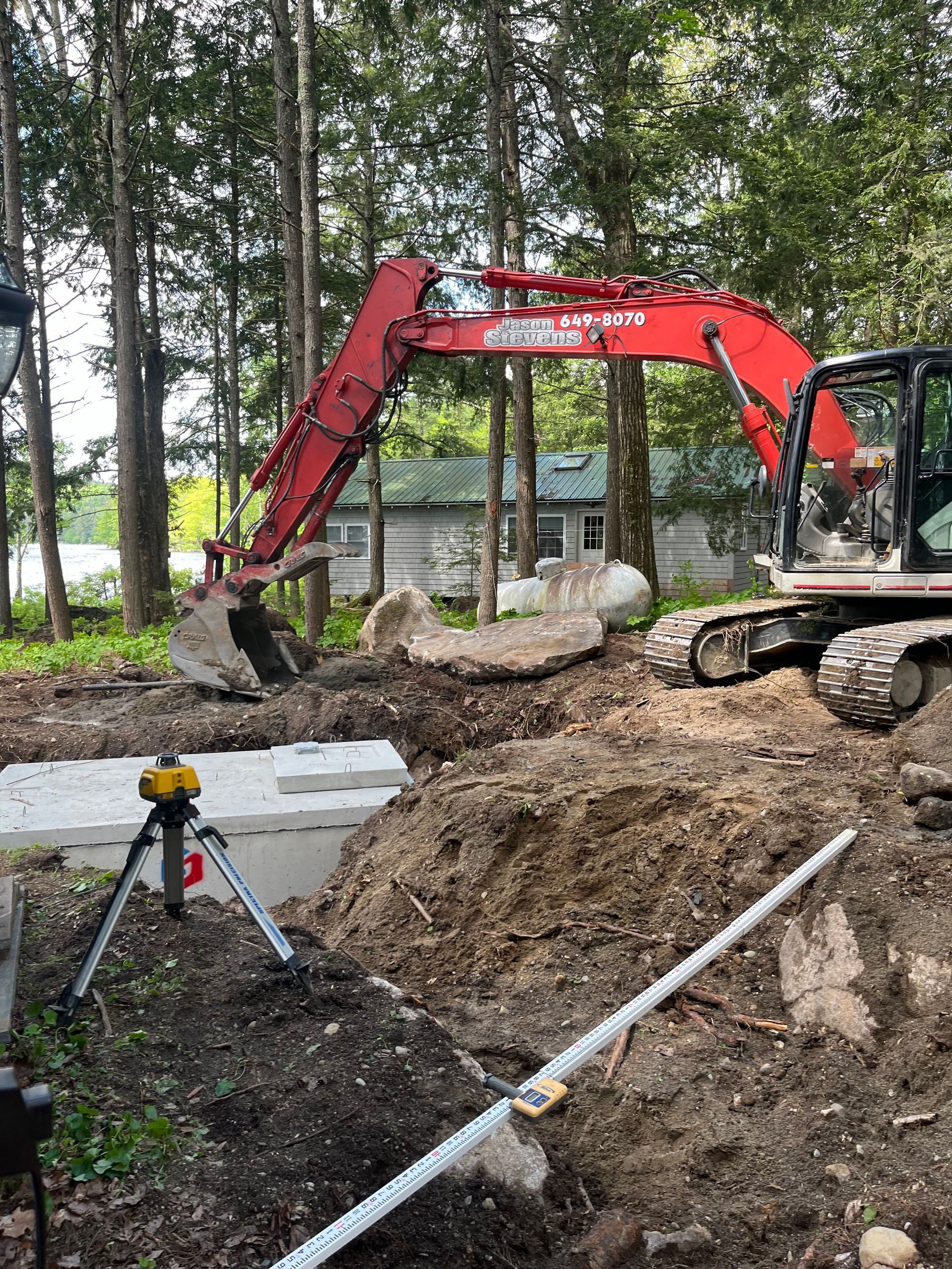 A red excavator operates at a construction site with a concrete septic tank and a surveying laser level in the foreground.