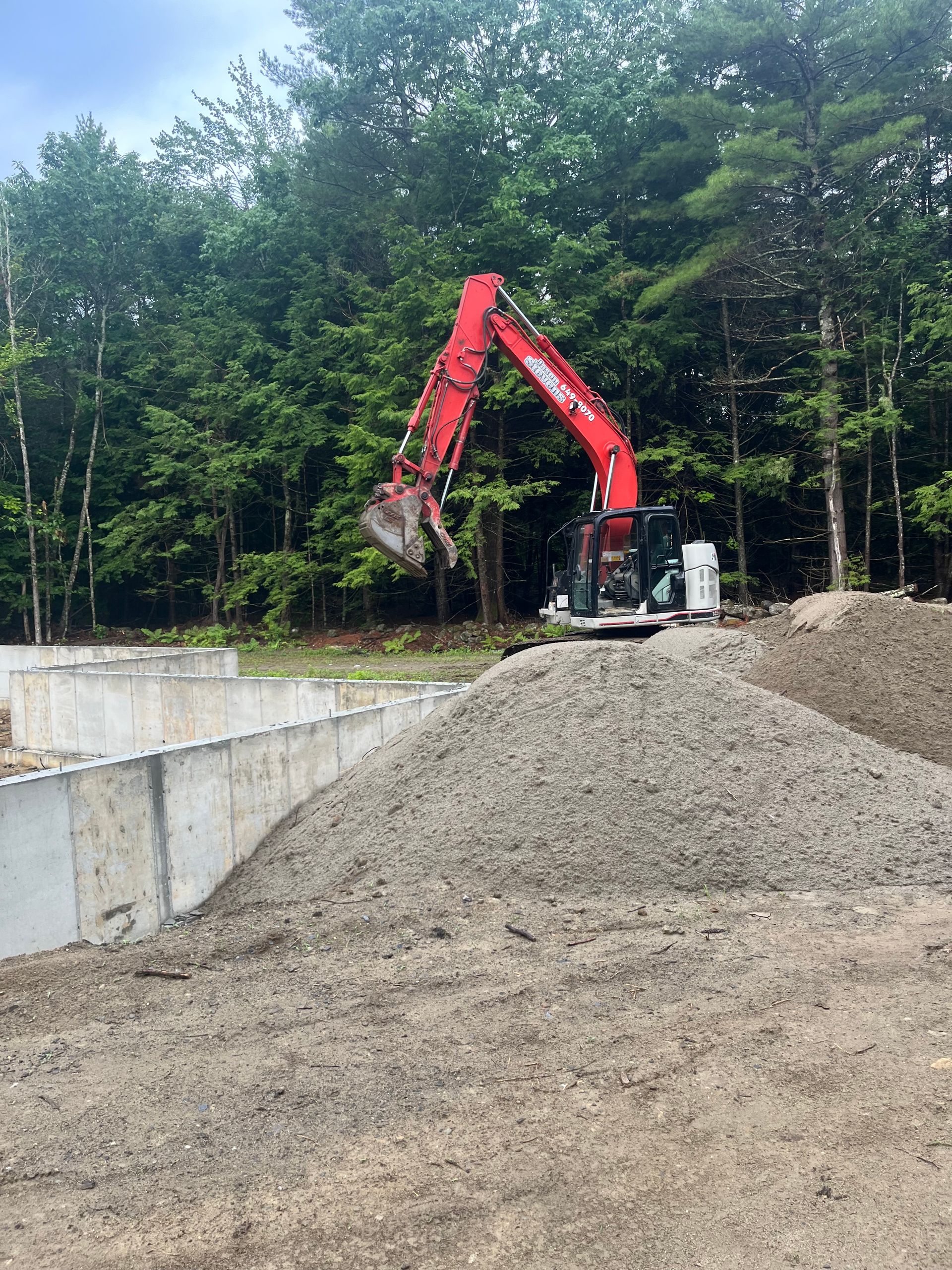 A red excavator sits atop a pile of gravel next to a concrete foundation at a construction site near a forest.