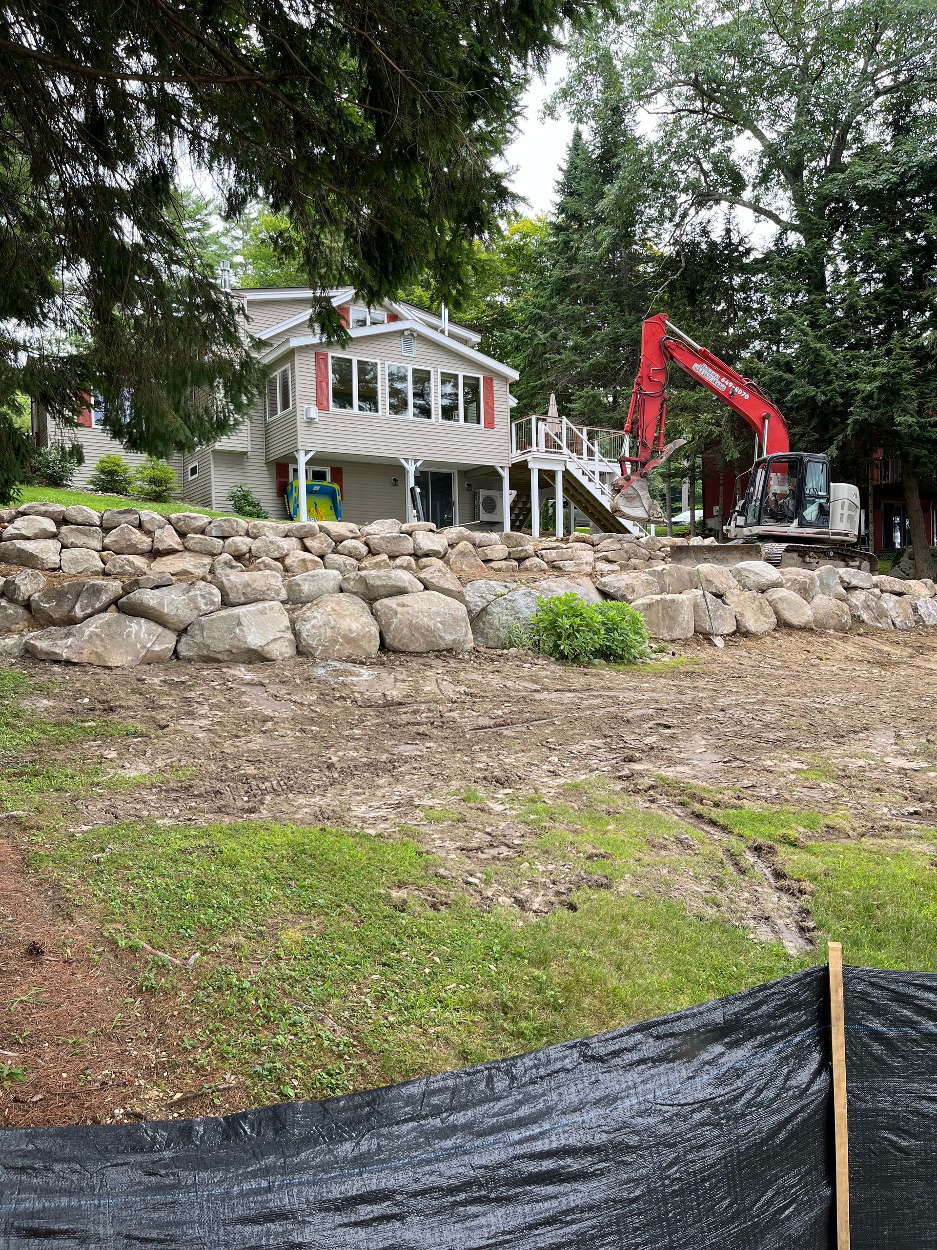A red excavator sits near a multi-story house set behind a large, tiered stone retaining wall on a grassy lot.